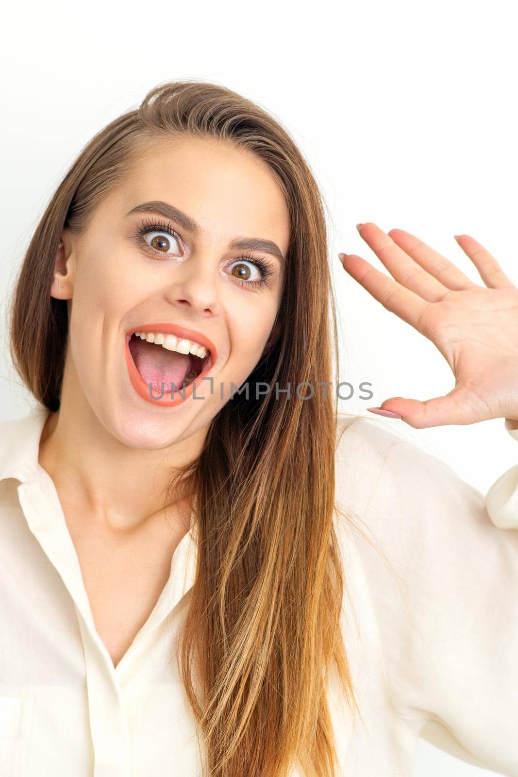 Portrait of young caucasian woman wearing white shirt raises hands and laughs positively with open mouth poses against a white background. by okskukuruza