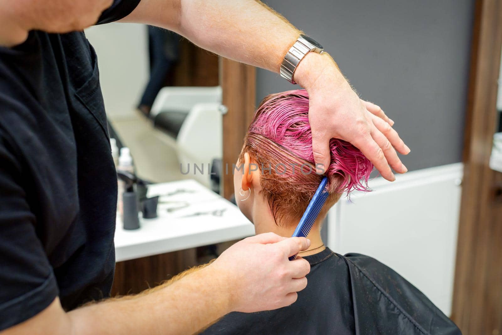 A hairdresser is combing the dyed pink wet short hair of the female client in the hairdresser salon, back view