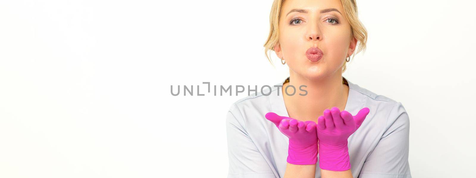 Young Caucasian doctor woman wearing pink gloves folding lips and holding palms to send air kiss isolated white background