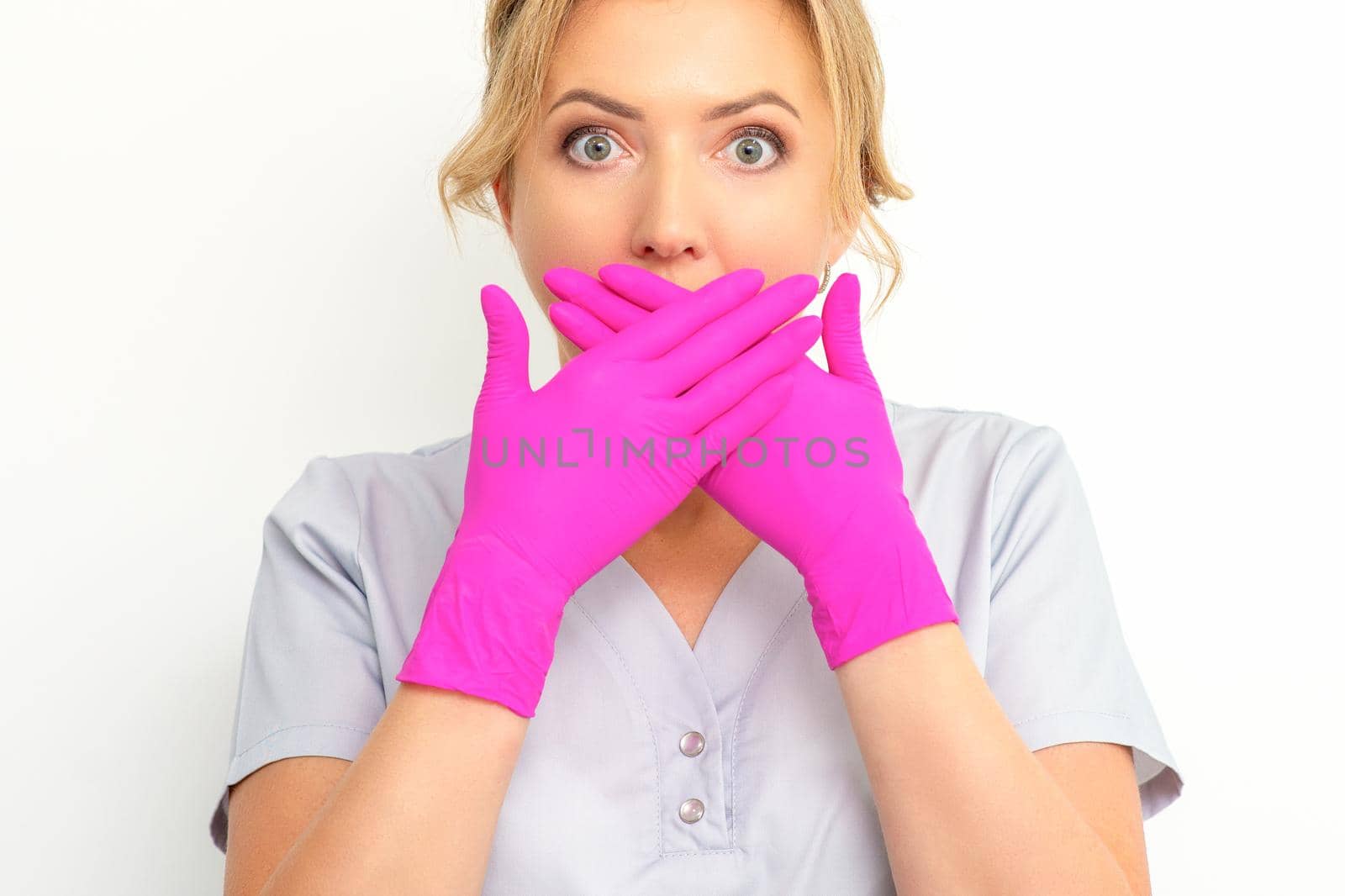Portrait of a young female caucasian doctor or nurse is shocked covering her mouth with her pink gloved hands against a white background
