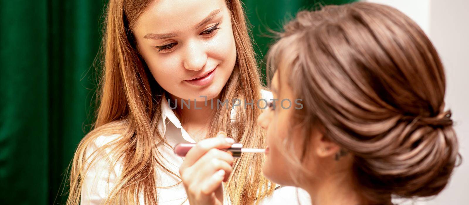 Makeup in the process. The makeup artist applies pink gloss lipstick on the lips of the beautiful face of the young caucasian woman in a beauty salon