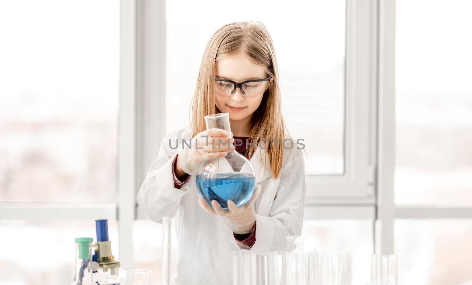 Smart girl doing scientific chemistry experiment wearing protection glasses, holding bottle and measuring blue liquid. Schoolgirl with chemical equipment on school lesson