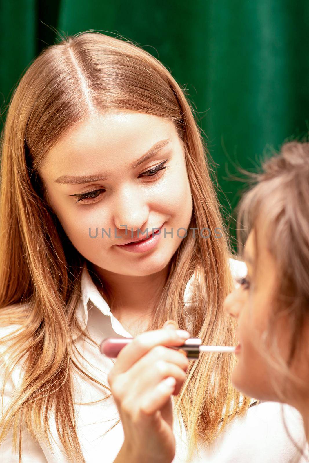 Makeup in the process. The makeup artist applies pink gloss lipstick on the lips of the beautiful face of the young caucasian woman in a beauty salon