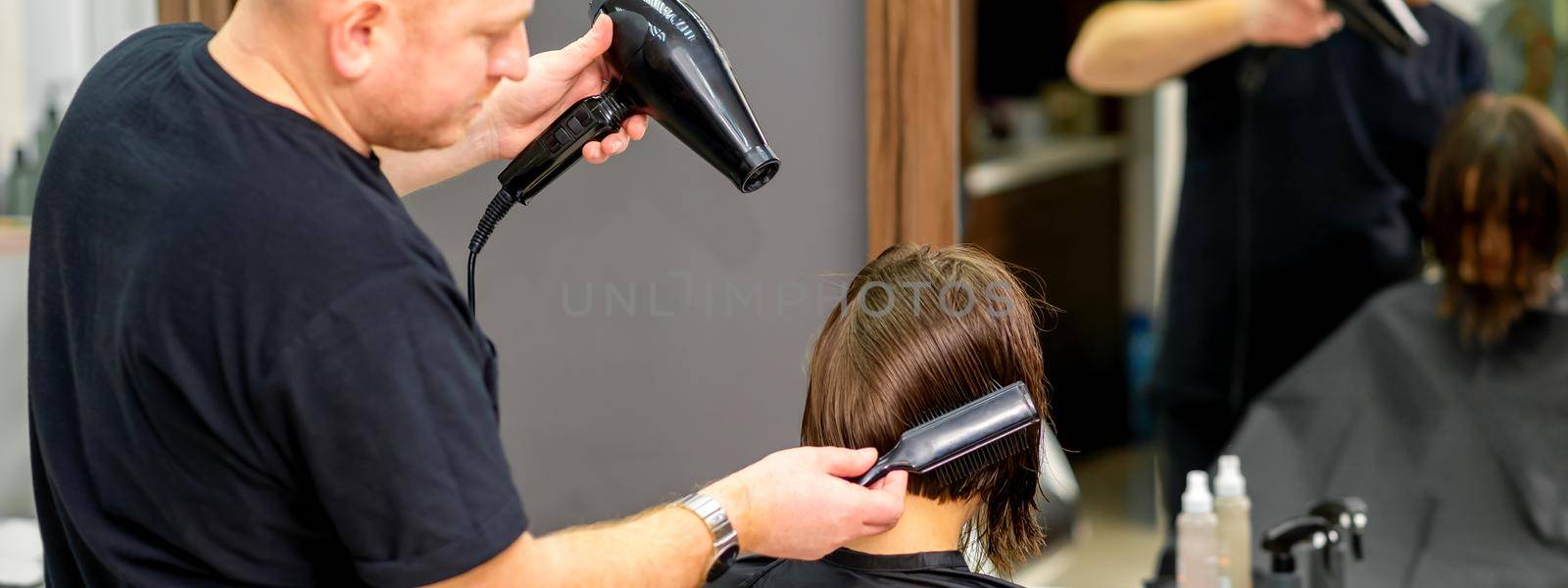 Male hairdresser drying short hair of young caucasian brunette woman with a black hairdryer and black round brush in a hairdresser salon