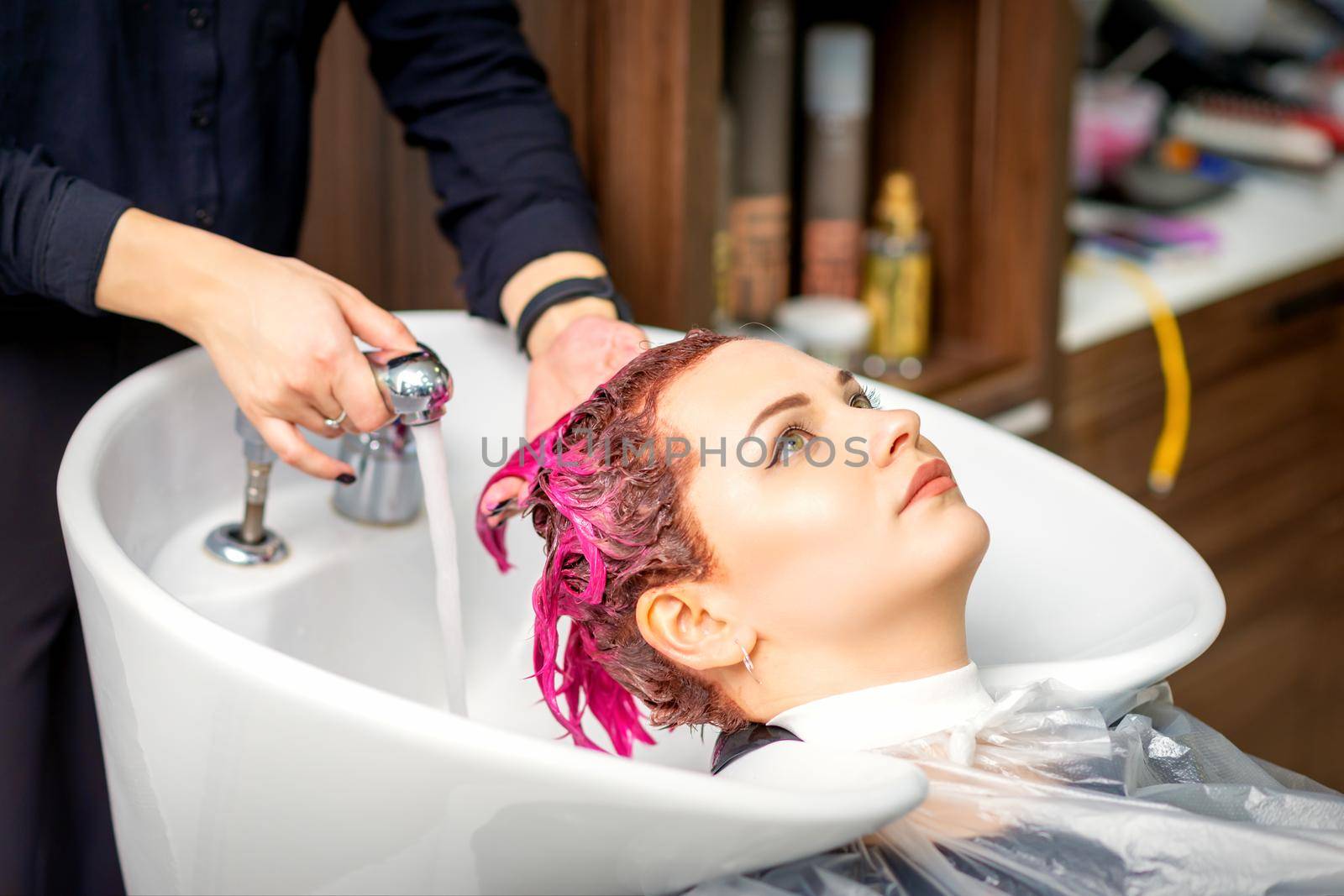Washing dyed female hair. A young caucasian woman having her hair washed in a beauty salon. Professional hairdresser washes pink color paint off of a customer's hair