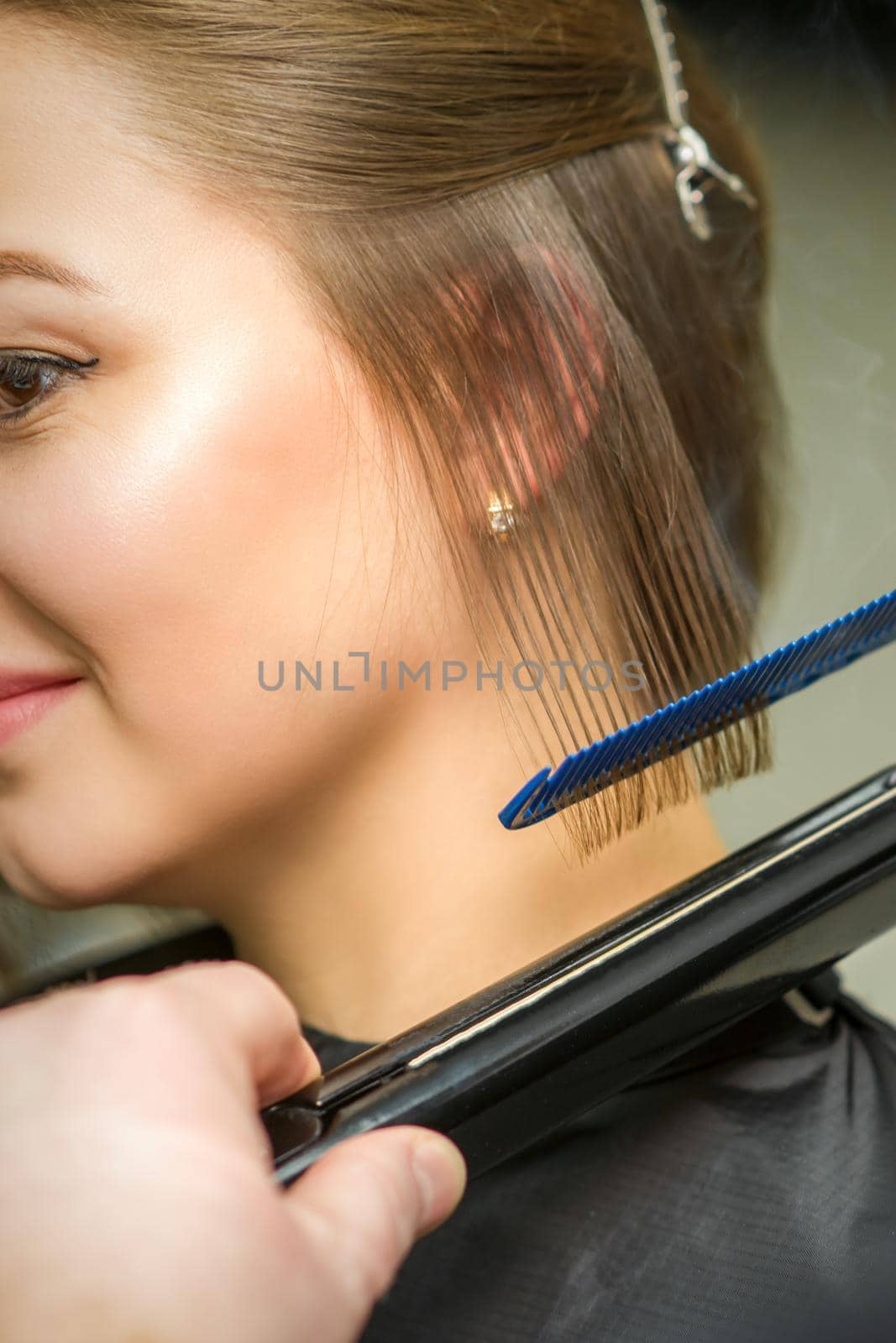 Hairstylist is straightening short hair of young brunette woman with a flat iron in a hairdresser salon, close up