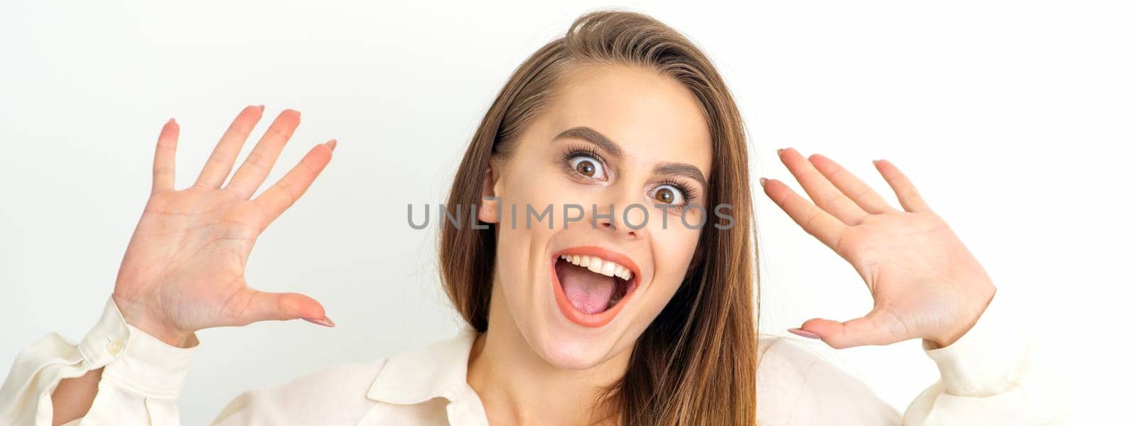 Portrait of young caucasian woman wearing white shirt raises hands and laughs positively with open mouth poses against a white background. by okskukuruza