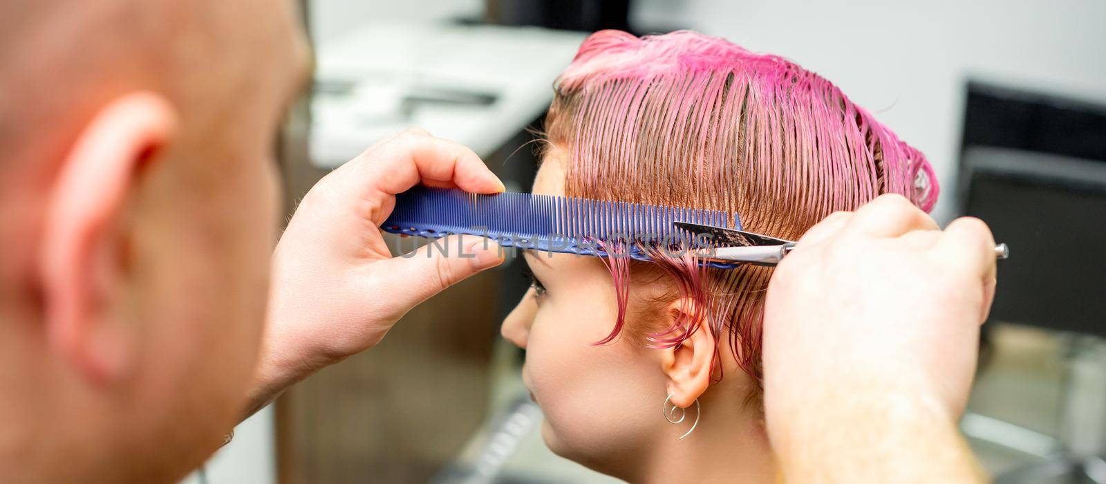 Haircut of dyed short pink wet hair of young caucasian woman by a male hairdresser in a barbershop