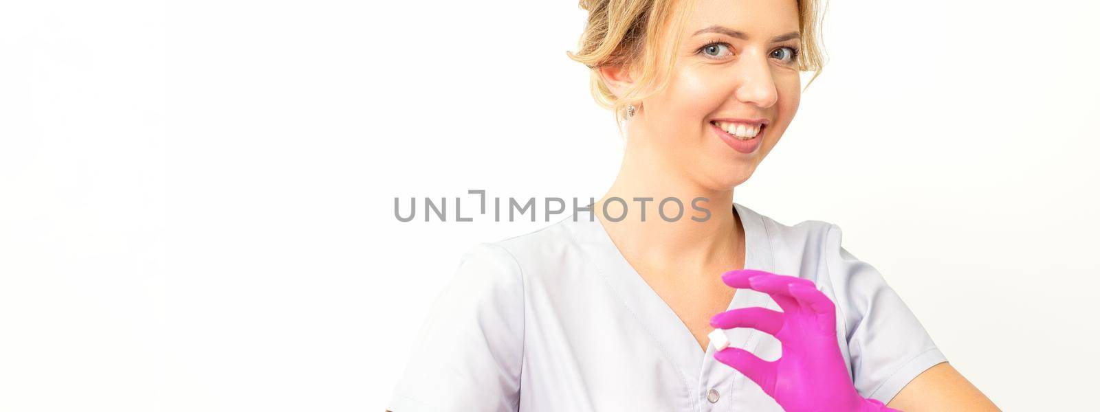 Portrait of a young smiling Caucasian beautician wearing pink gloves holding sugar cubes showing and looking at the camera against a white background. by okskukuruza