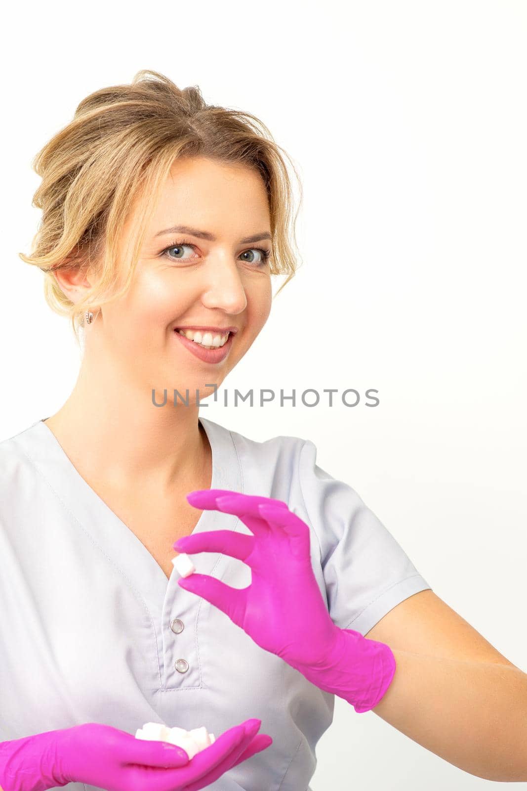 Portrait of a young smiling Caucasian beautician wearing pink gloves holding sugar cubes showing and looking at the camera against a white background. by okskukuruza