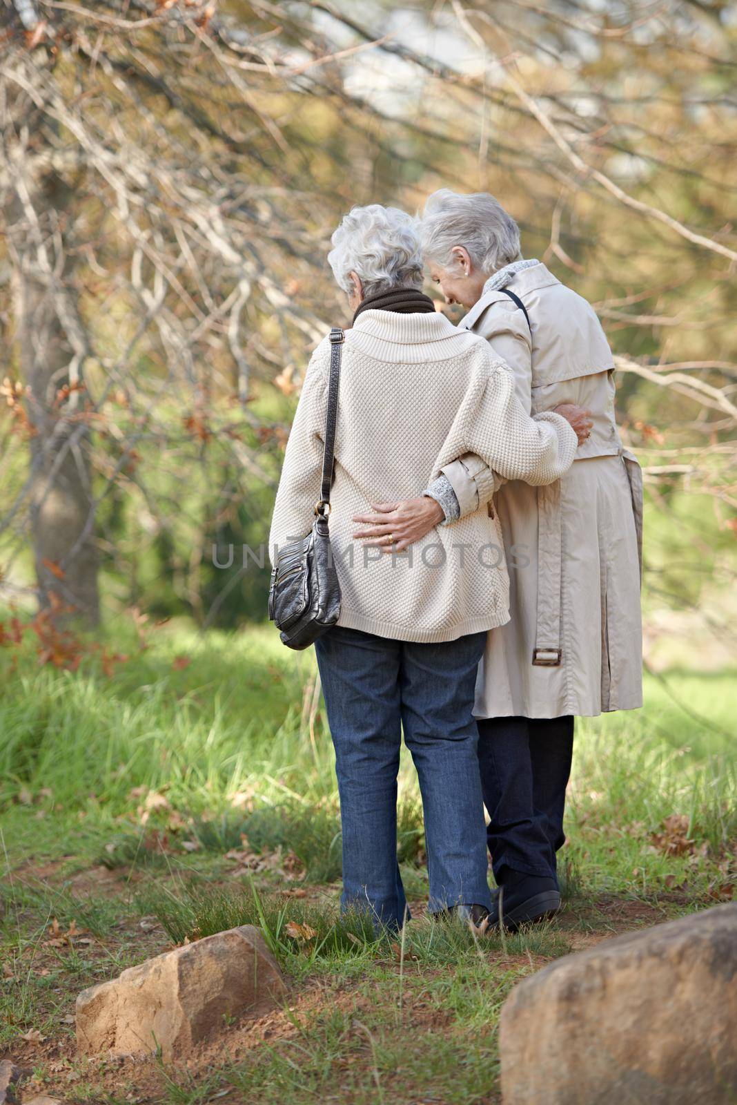 They are each others support. Two senior ladies embracing with autumn shaded trees in the background. by YuriArcurs