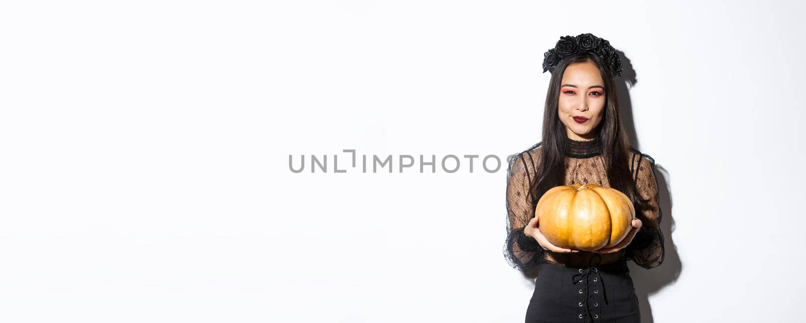 Image of cunning asian woman in black dress, impersonating evil witch on halloween, holding big pumpkin, standing over white background.