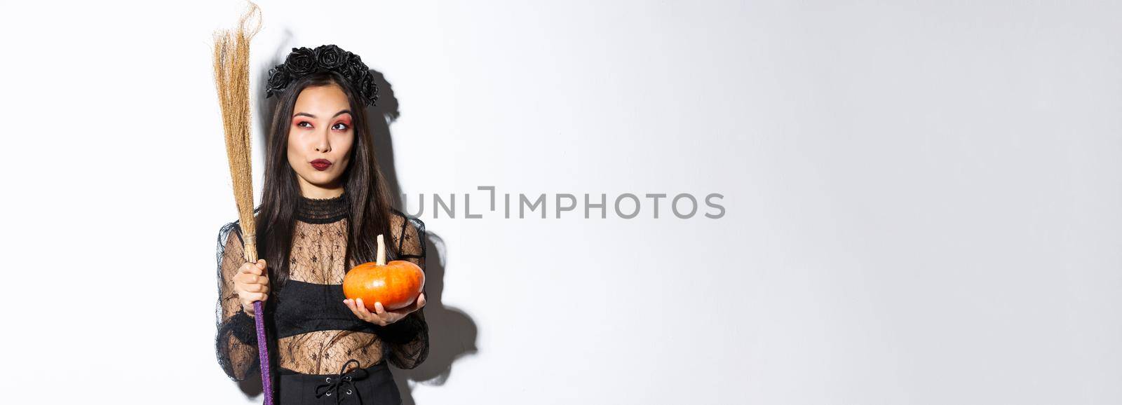 Image of sassy witch in gothic lace dress, holding broom and pumpkin, looking at upper left corner with halloween banner, white background.