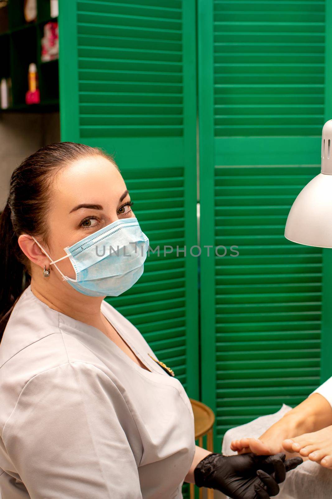 Young female podiatrist with a protective mask doing chiropody and looking at the camera in her podiatry clinic