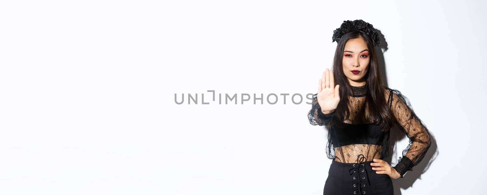 Confident elegant young witch showing stop gesture, woman dressed-up in halloween costume extend one hand to prohibit something, express disapproval, standing white background.