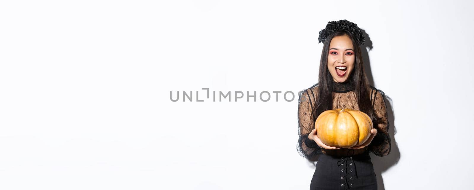 Image of joyful asian woman in witch costume, celebrating halloween, giving you pumpkin and looking scary, standing over white background by Benzoix