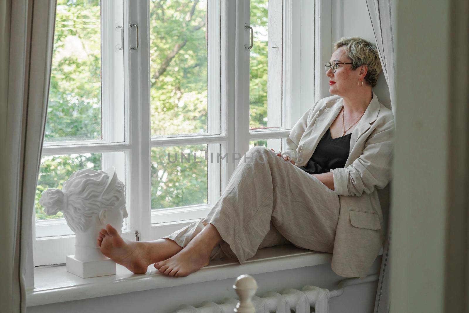A middle-aged woman in a beige suit and black tank top sits mysteriously and looks out the window on the windowsill. Green trees outside.