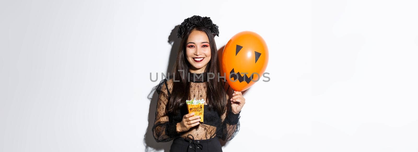Image of cheerful asian girl trick or treating on halloween, holding sweets and orange balloon with scary face.