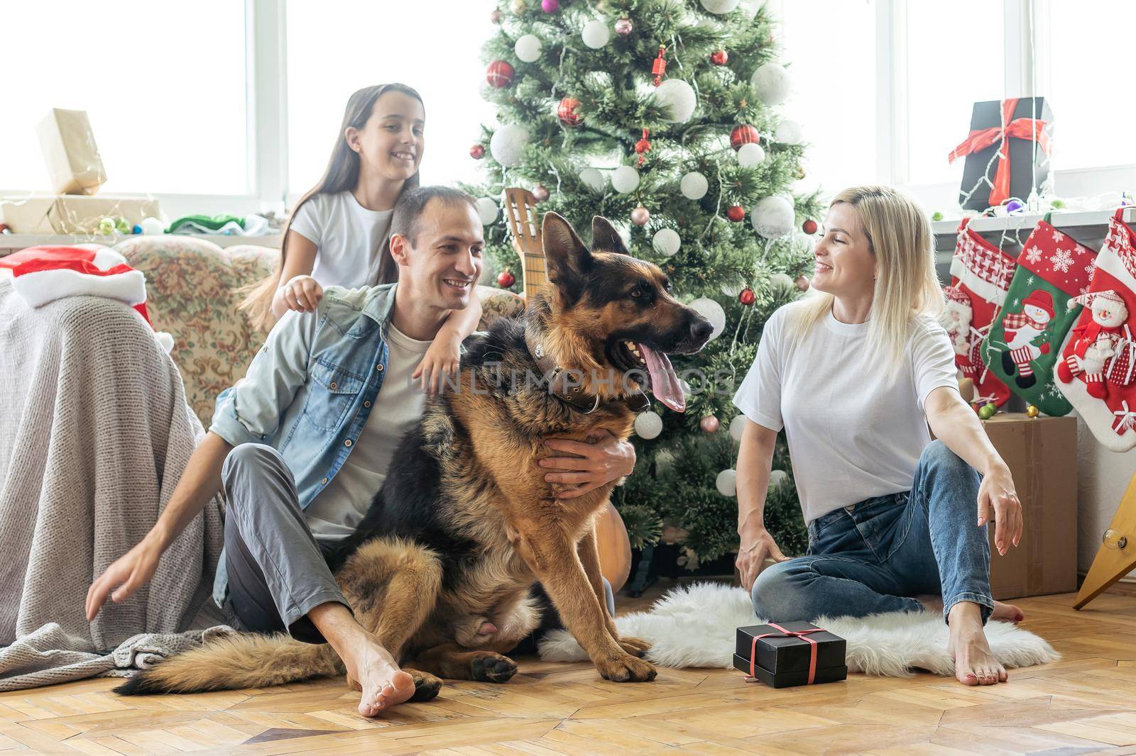 Friendly family is playing with dog near Christmas tree. They are sitting and laughing by Andelov13