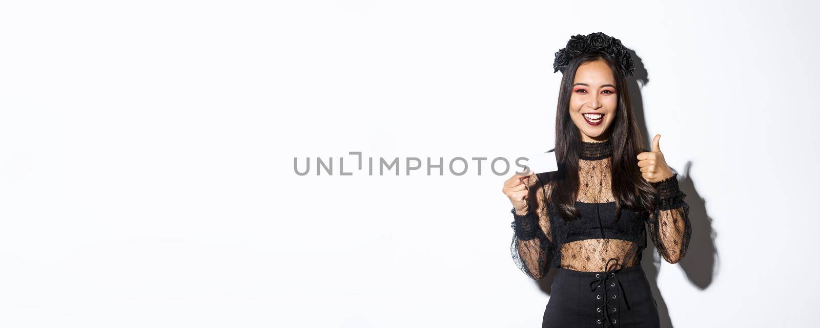 Portrait of satisfied asian woman in elegant lace dress and black wreath showing thumbs-up in approval and demonstrate her credit card, standing over white background by Benzoix