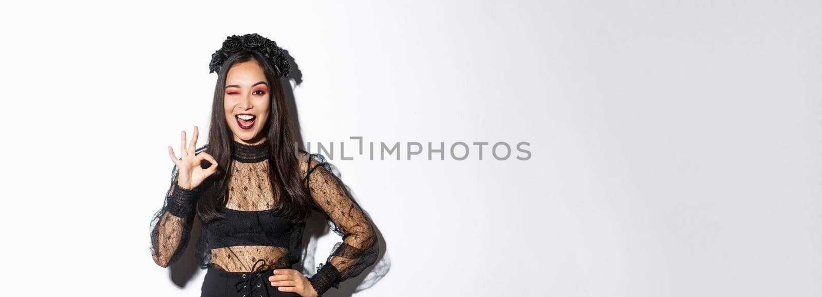 Attractive young asian woman in halloween gothic dress, showing okay gesture, give permission, approve or like your party costume, standing pleased over white background.