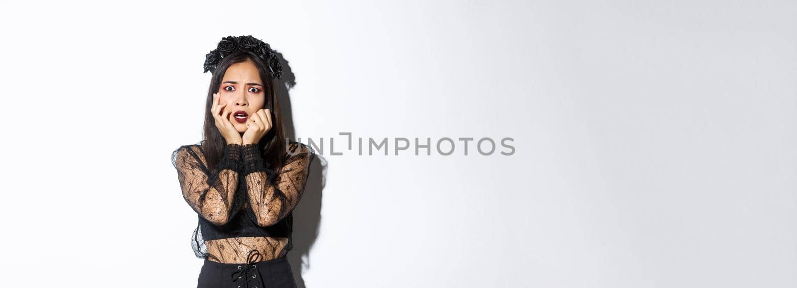 Image of scared and shocked asian female in halloween costume looking frightened at something scary, standing over white background.