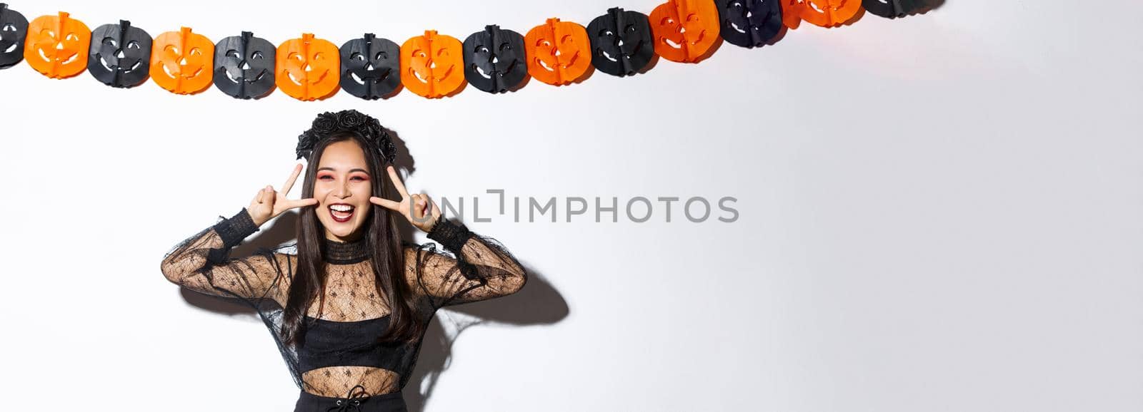 Cute happy asian woman enjoying halloween party, showing peace gesture and smiling, wearing witch costume, standing against pumpkin banners decoration.
