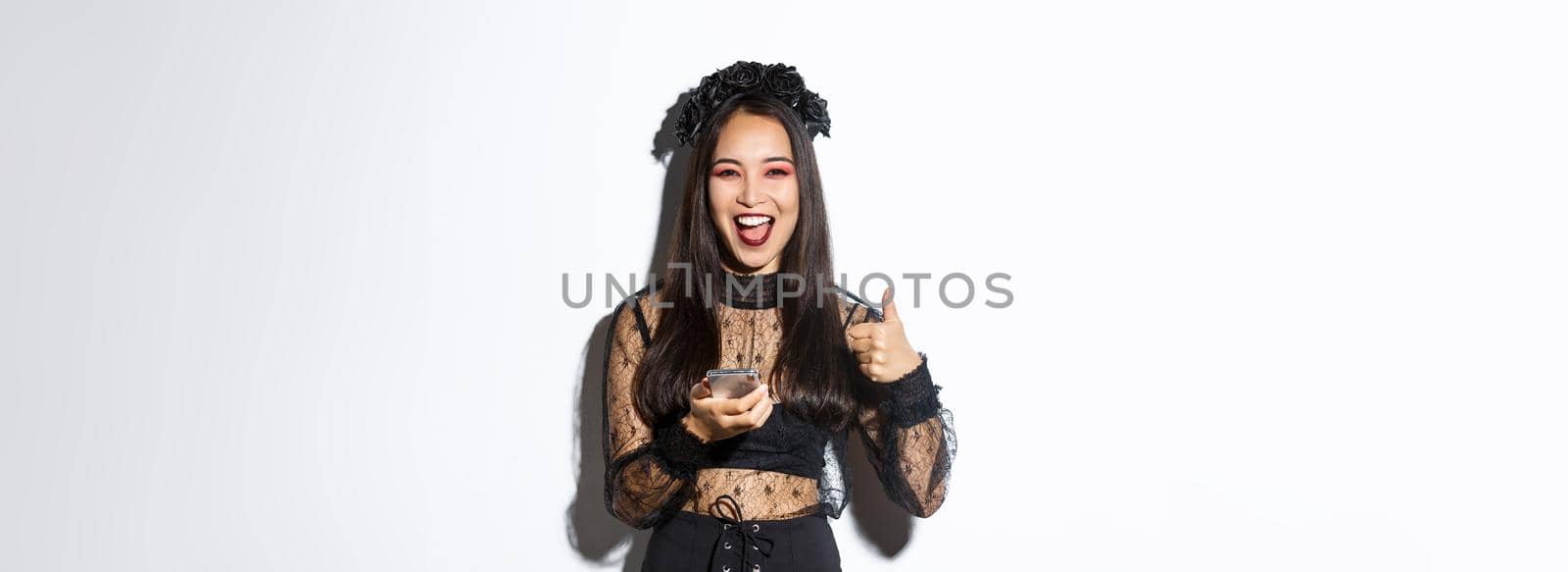 Portrait of satisfied asian woman in elegant gothic dress and black wreath showing thumbs-up while using mobile phone, standing over white background by Benzoix
