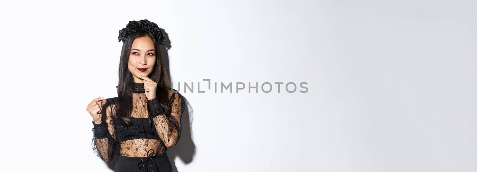 Image of smiling beautiful asian woman in gothic lace dress and wreath, thinking while holding credit card, standing over white background.