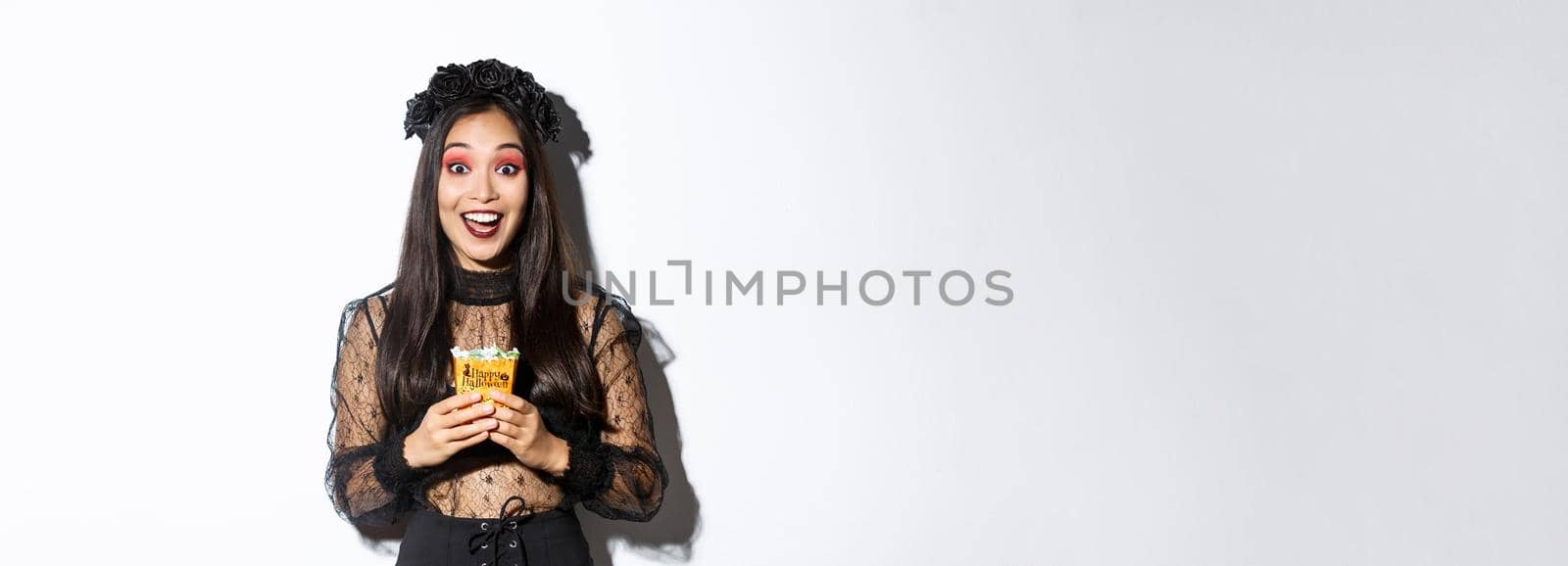 Beautiful asian girl smiling happy, holding sweets, wearing witch costume on halloween, enjoying trick or treating.