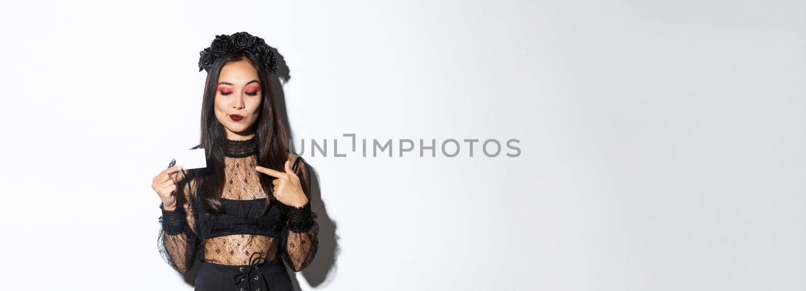 Image of curious beautiful asian girl in witch costume pointing finger at credit card, standing over white background.