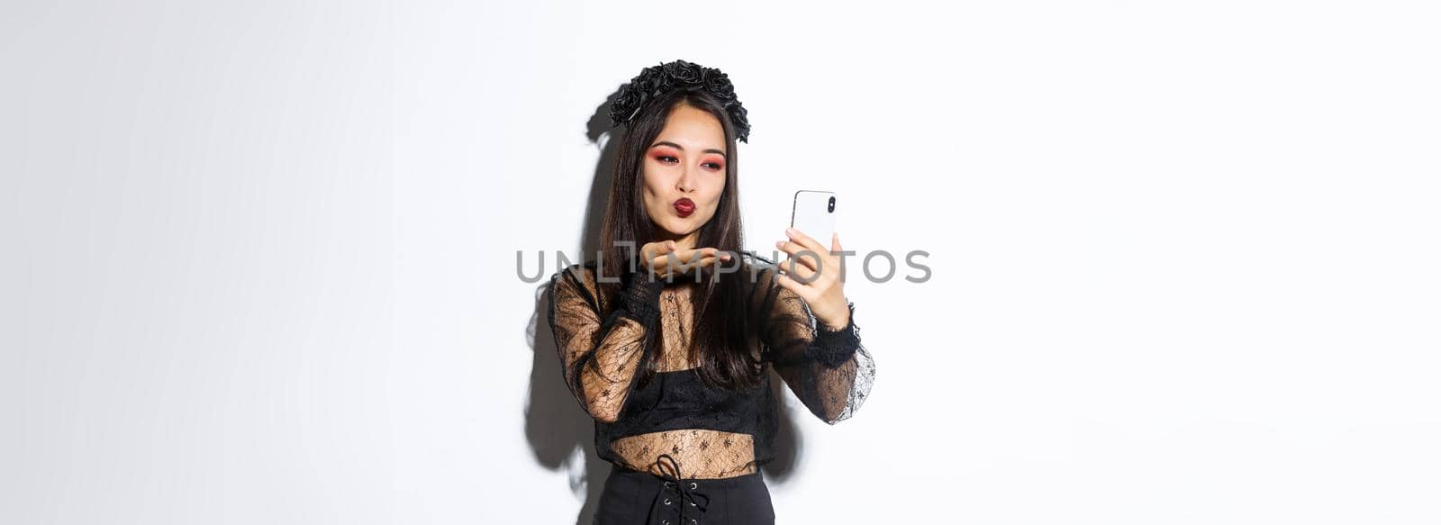 Portrait of stylish asian female blogger with gothic makeup and halloween costume sending air kiss at mobile phone camera, record video or having videocall, standing over white background.