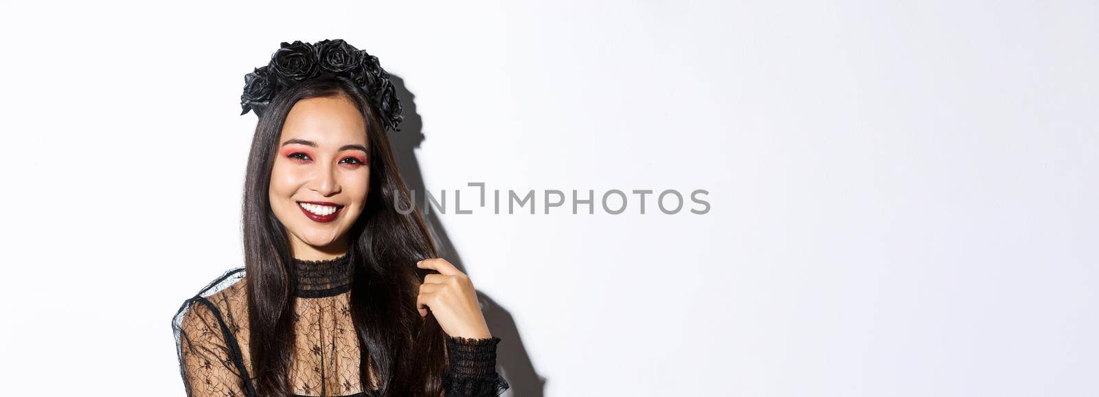 Close-up of beautiful elegant asian woman in black wreath and gothic lace dress smiling, standing over white background, dressed-up for halloween party.