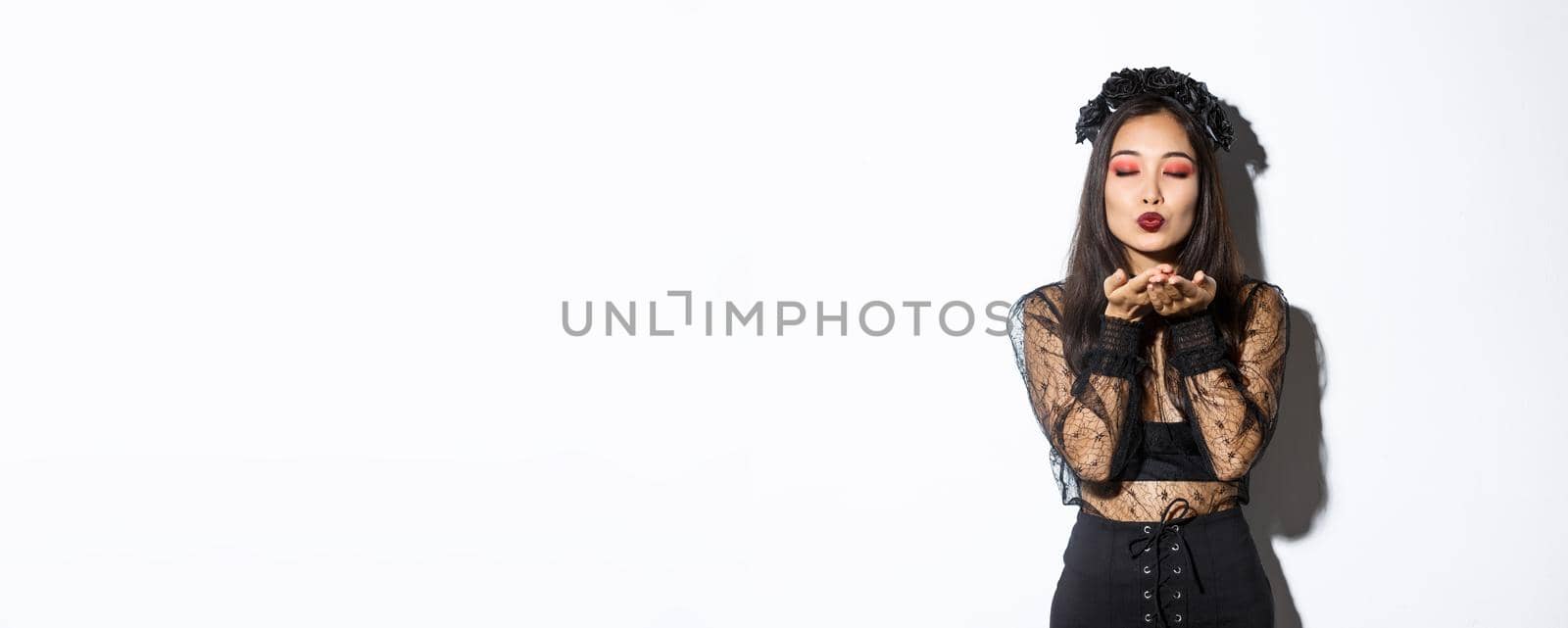 Image of beautiful and elegant asian woman in witch costume, close eyes and sending air kiss t camera, wearing gothic dress with black wreath, standing over white background.