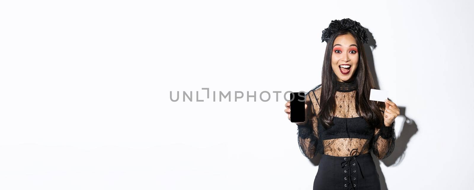 Cheerful beautiful asian woman in halloween costume, dressed as witch and showing smartphone with credit card, standing over white background.