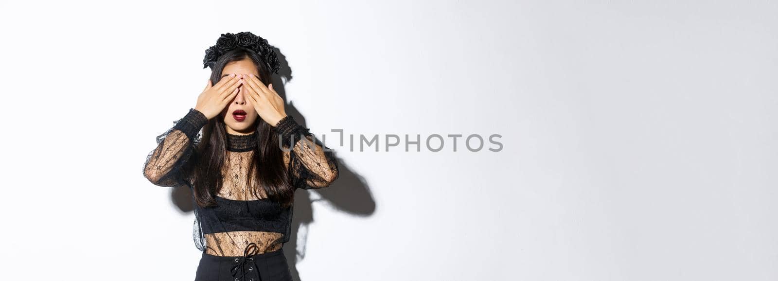 Hopeful and excited stylish asian girl in witch dress close her eyes and waiting for surprise, open mouth amused, wearing halloween costume, standing over white background.