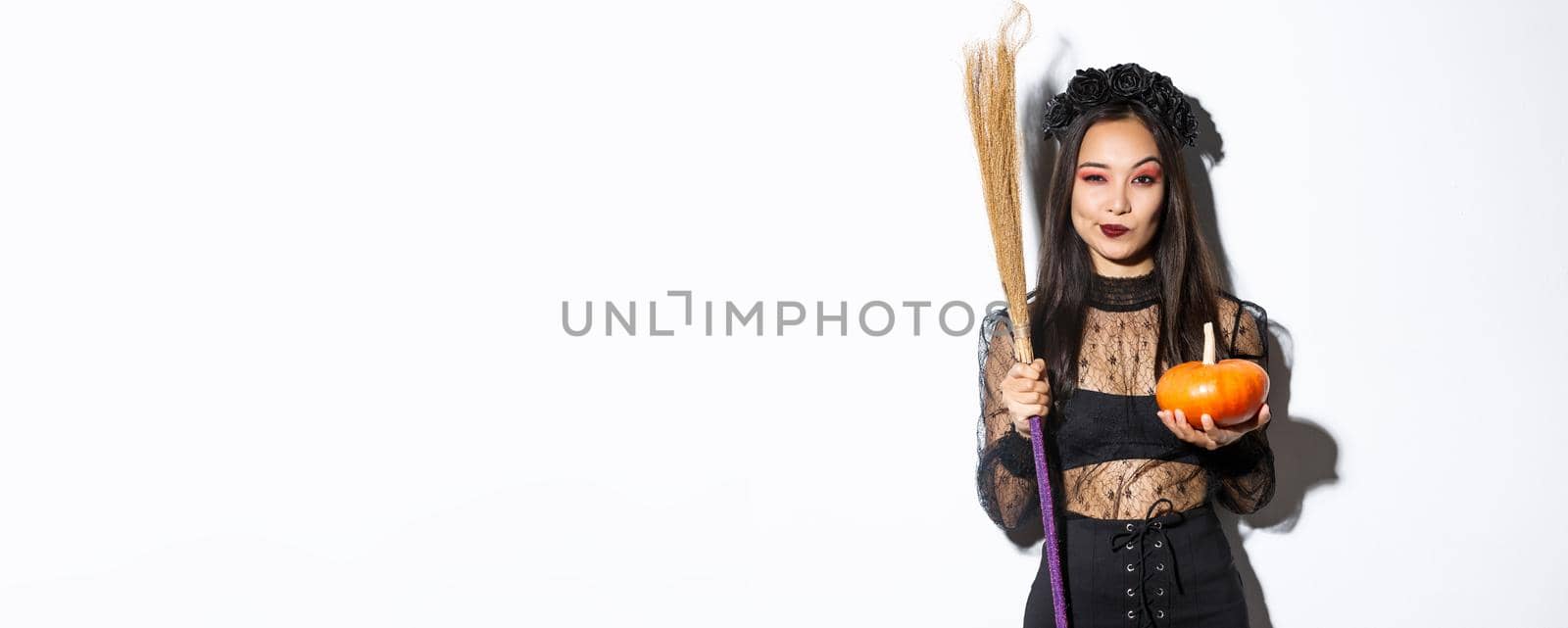 Image of beautiful asian woman dressed-up as a witch for halloween party, holding broom and pumpkin, standing over white background.