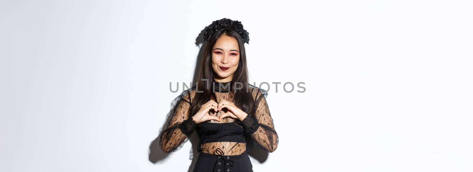 Image of lovely smiling asian woman love celebrating halloween, showing heart gesture and looking at camera, standing over white background in gothic lace dress with wreath.