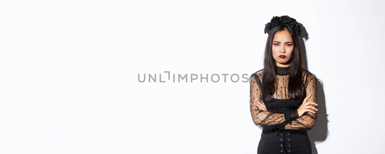 Image of angry and offended asian girl complaining on something, cross arms and sulking, looking insulted or jealous, standing over white background in halloween party dress.
