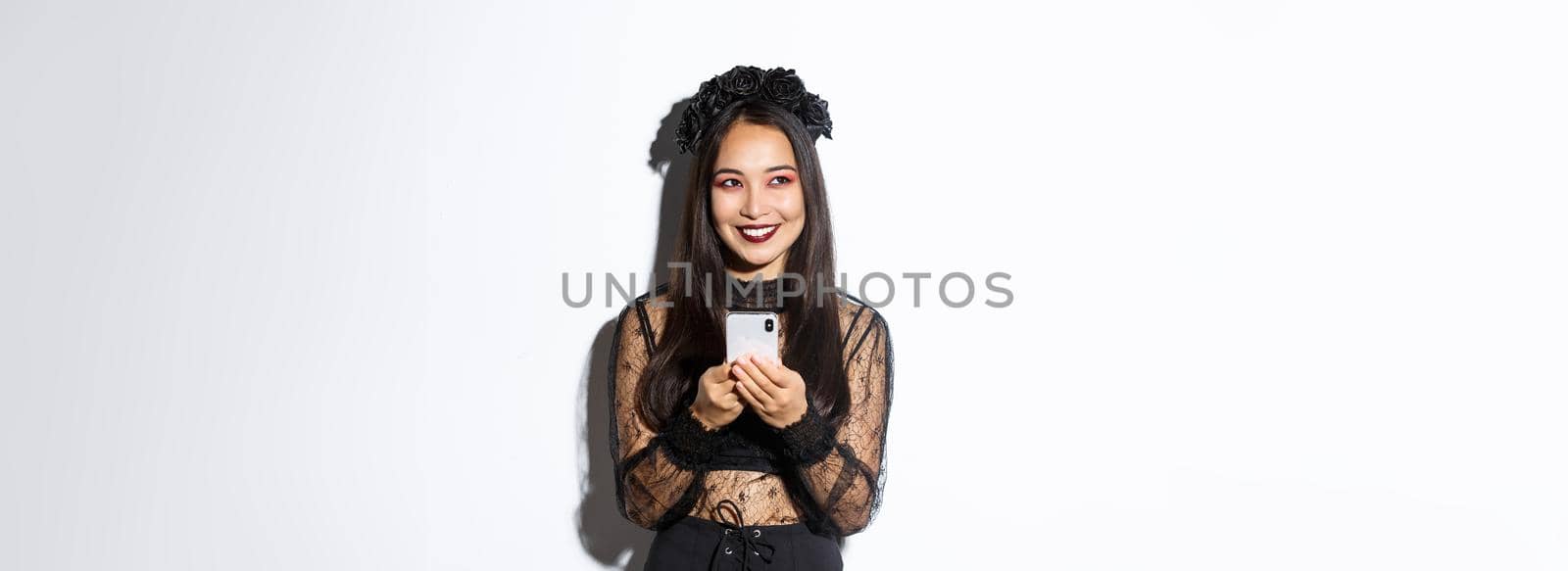 Portrait of cunning beautiful asian woman, witch in gothic lace dress using mobile phone, smiling and looking at upper left corner, standing over white background.