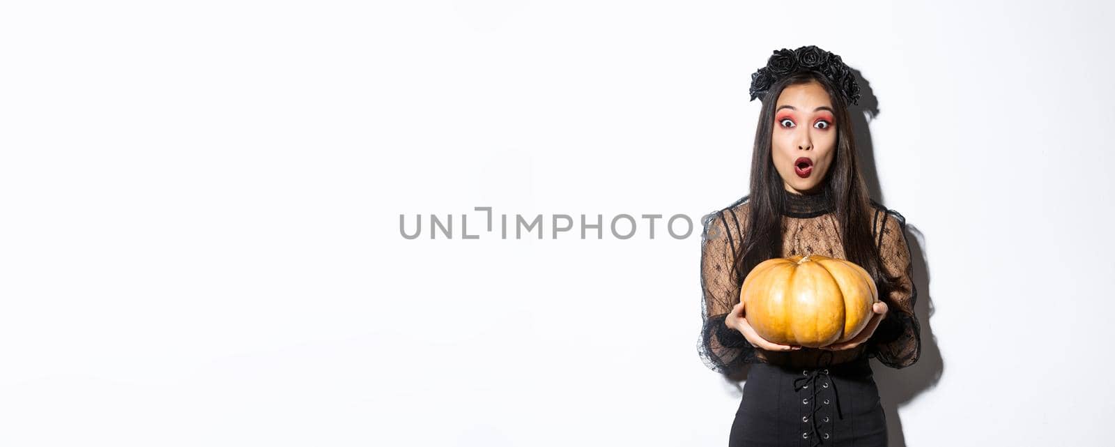 Surprised asian girl in witch costume, holding pumpkin and gasping amazed at camera, preparing for halloween holiday, standing over white background.