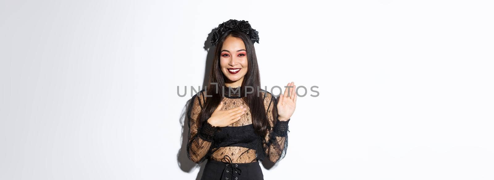 Image of cute sincere asian girl in halloween costume making promise, holding one hand on heart while swearing, being honest, standing over white background.