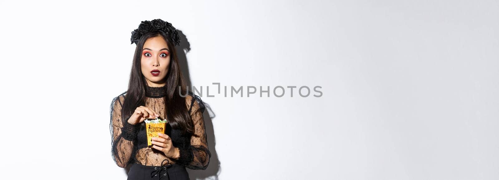 Image of surprised and fascinated asian woman in witch costume, holding sweets gathered during trick or treating, standing over white background.