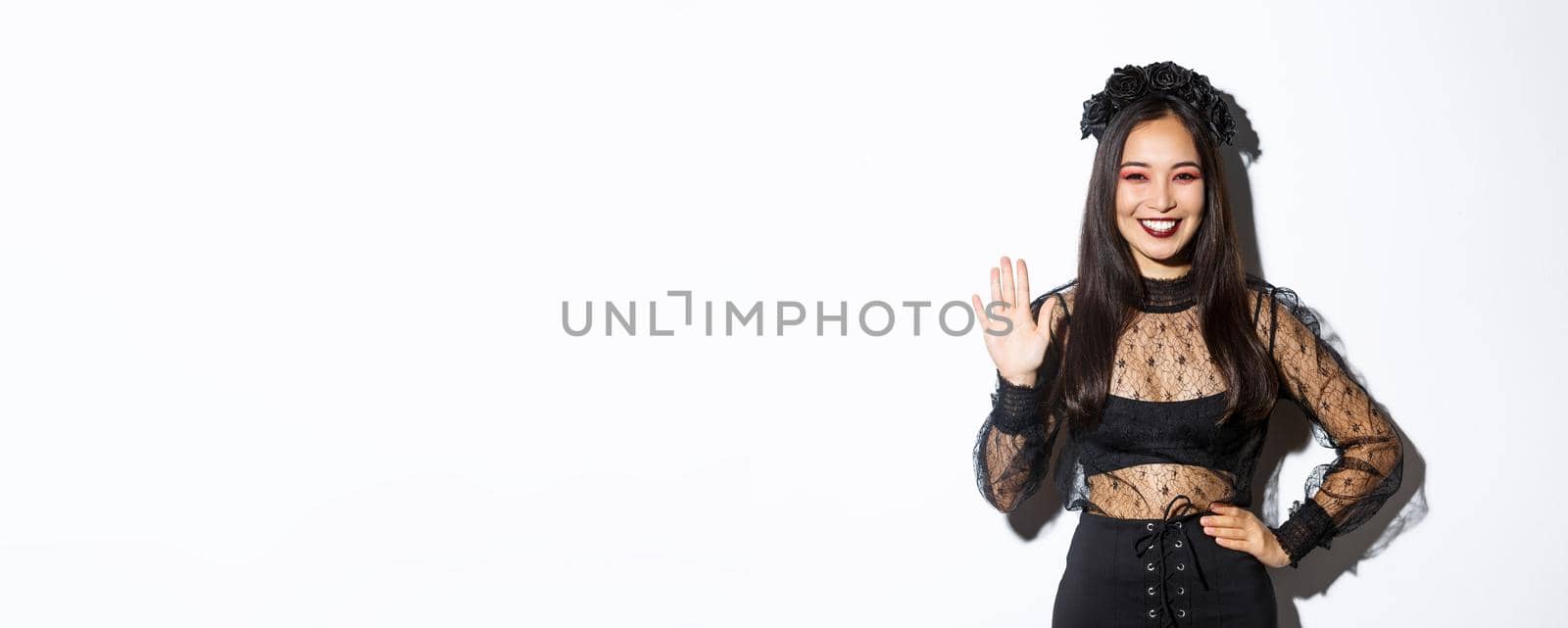 Image of friendly stylish asian woman in gothic lace dress waving hand to say hello, greeting someone, welcome people at halloween party, standing over white background.