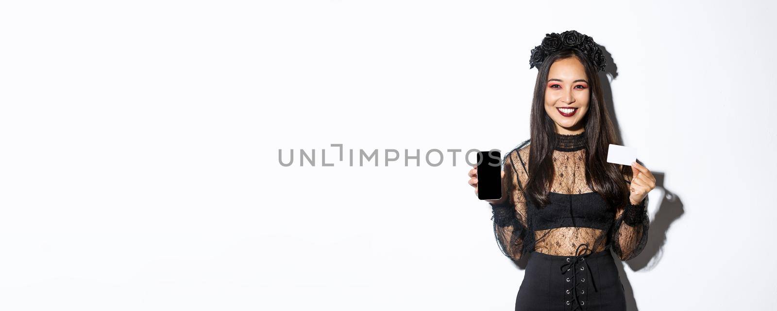 Smiling attractive woman in black gothic dress showing credit card and smartphone display, standing over white background in halloween costume by Benzoix