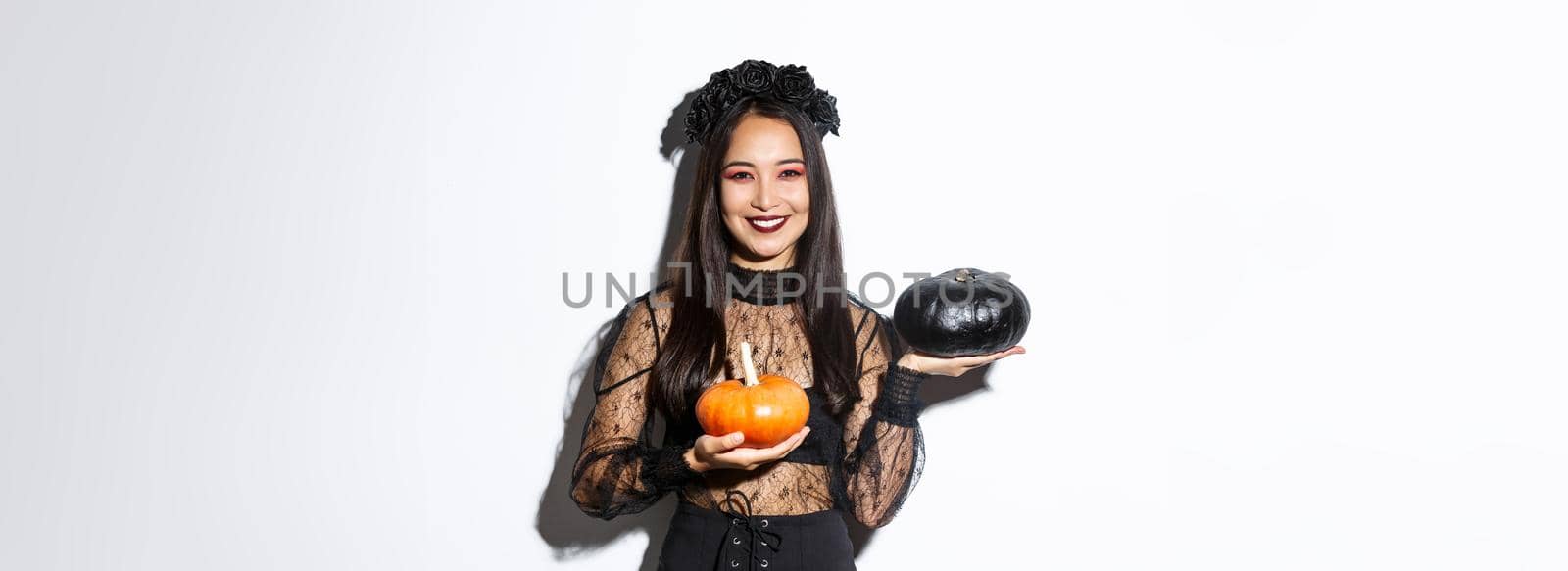 Image of attractive korean girl celebrating halloween in gothic lace dress, impersonating witch and holding two pumpkins.