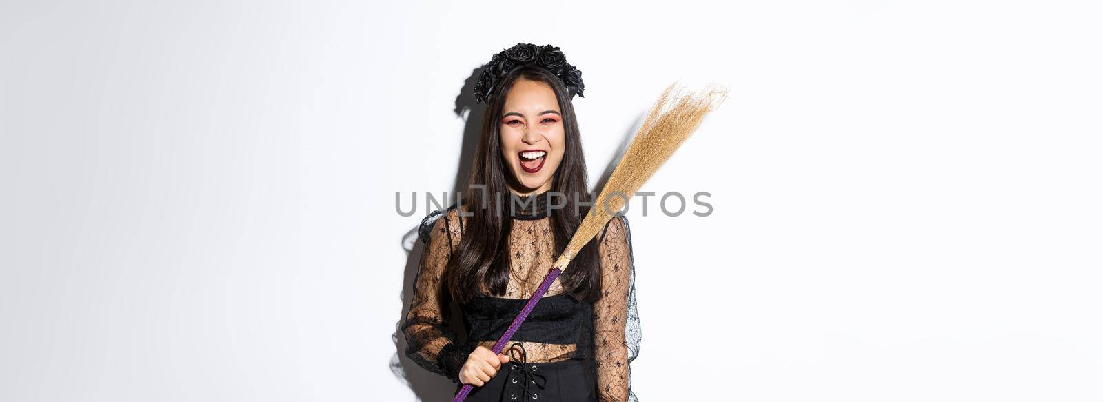 Sassy evil witch laughing and waving her broom, wearing halloween costume, standing over white background.