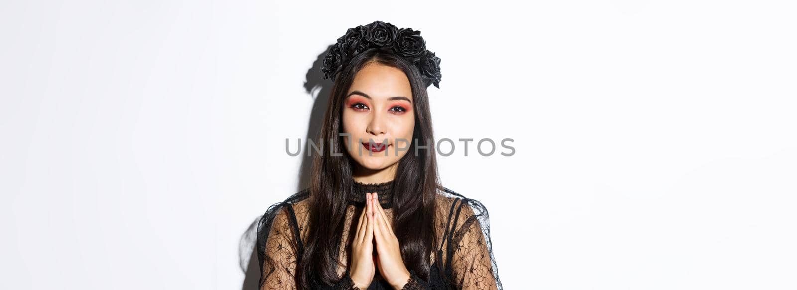 Close-up of young asian woman in black gothic dress and wreath holding hands in pray, girl wearing witch costume and celebrating halloween.