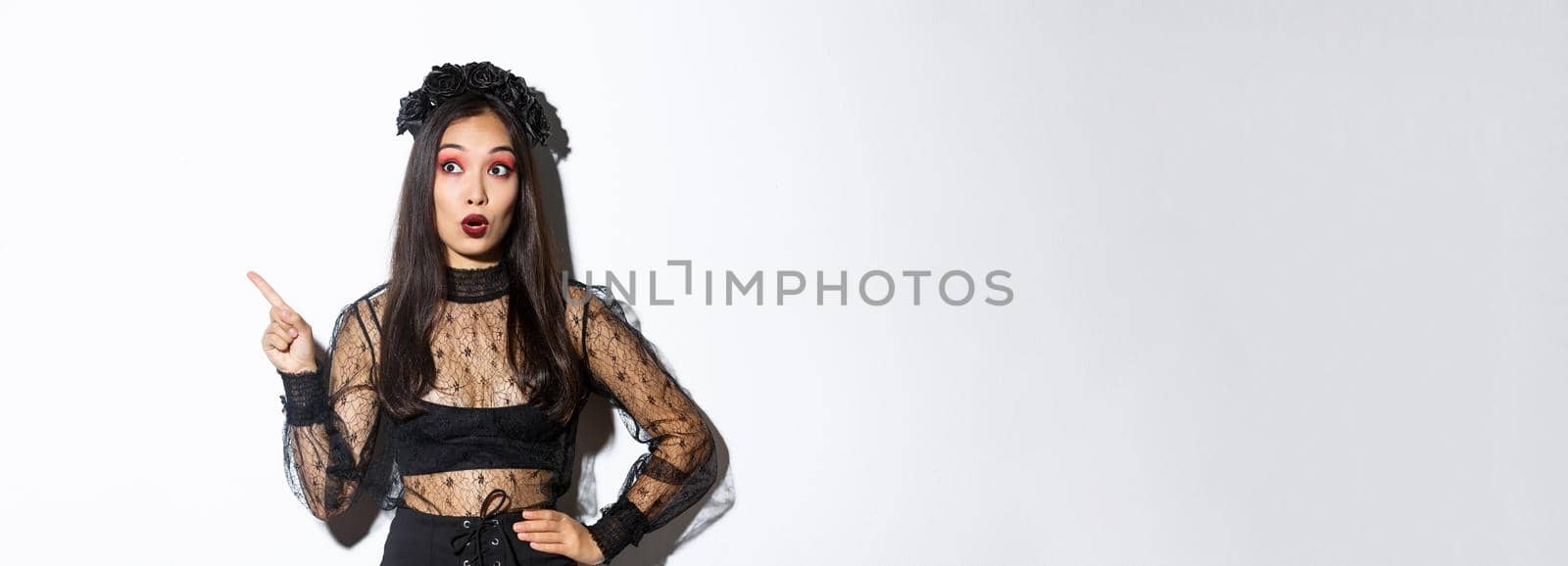 Intrigued asian girl celebrating halloween in black gothic dress and wreath, looking and pointing finger upper left corner at something interesting, standing over white background.