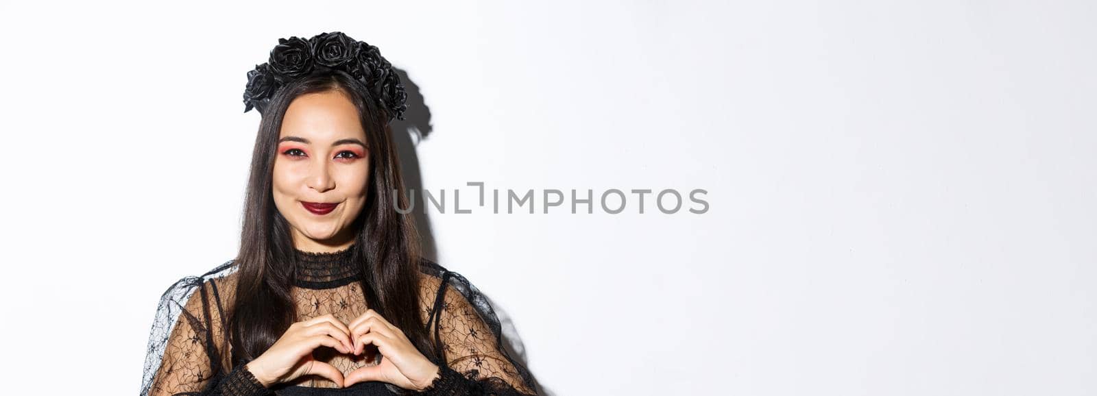 Close-up of smiling pretty asian woman in witch costume and black wreath showing heart gesture, loving halloween holiday, standing over white background.